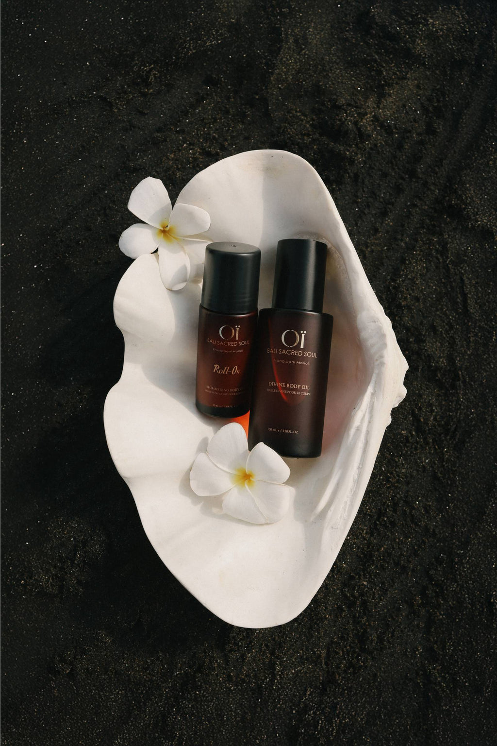 Flacons OÏ Divine et Roll-on dans un coquillage avec des fleurs de frangipanier sur une plage de sable noir